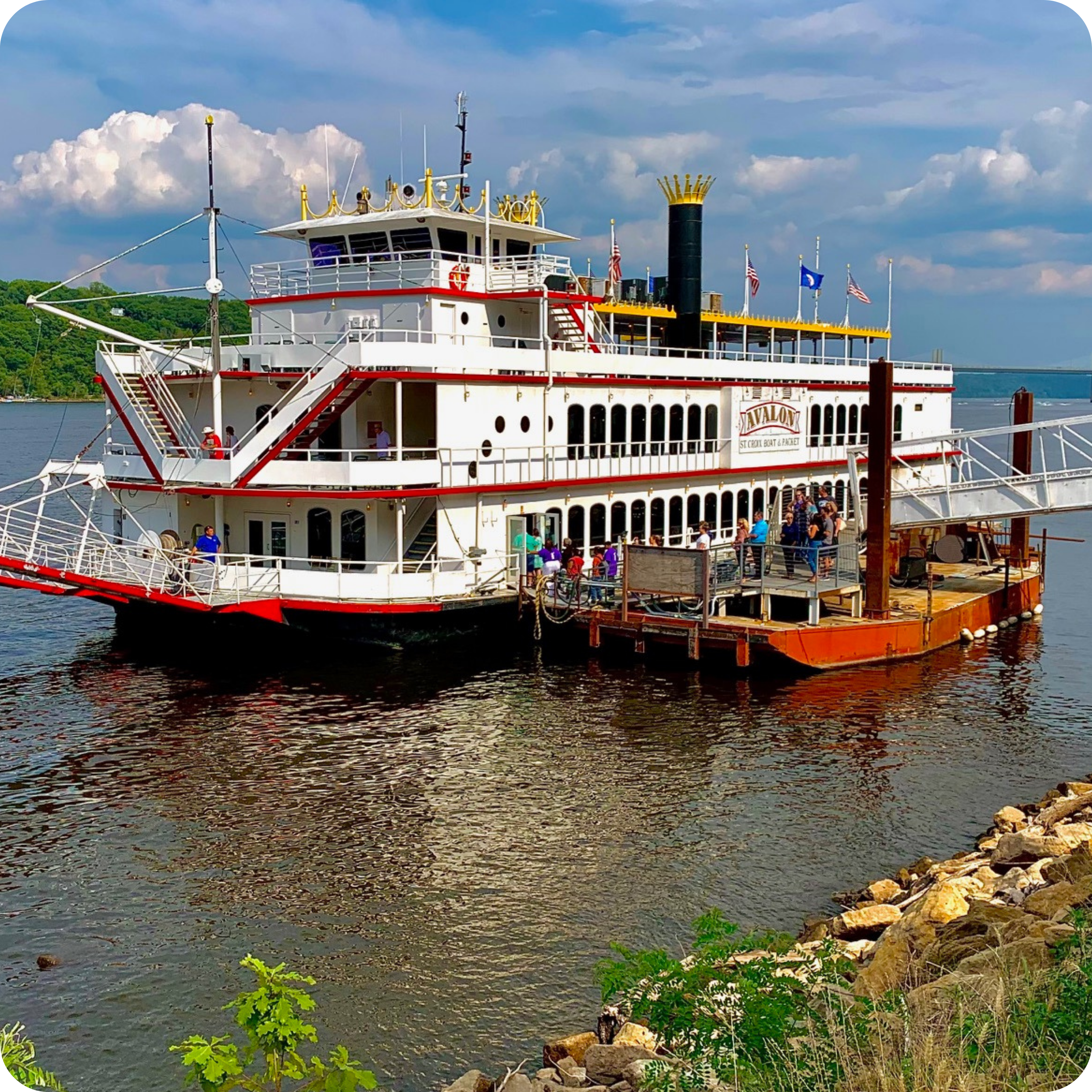 A large white and red riverboat, decorated with flags and a crowned smokestack, is docked alongside a pier, with people mingling on board and at the dock, set against a green hillside and a partly cloudy sky.